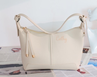 May include: Cream-coloured shoulder bag with a curved handle and a gold zip. The bag has the name "Cathy" embroidered on the front. The bag is sitting on a newspaper.