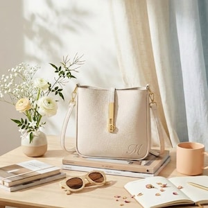 Puede incluir: Bolso de mano color crema con cierre y correa dorados. El bolso tiene forma rectangular con una textura sutil y una inicial "M" dorada en la parte delantera. El bolso está sobre una pila de libros, junto a gafas de sol y un jarrón con flores.