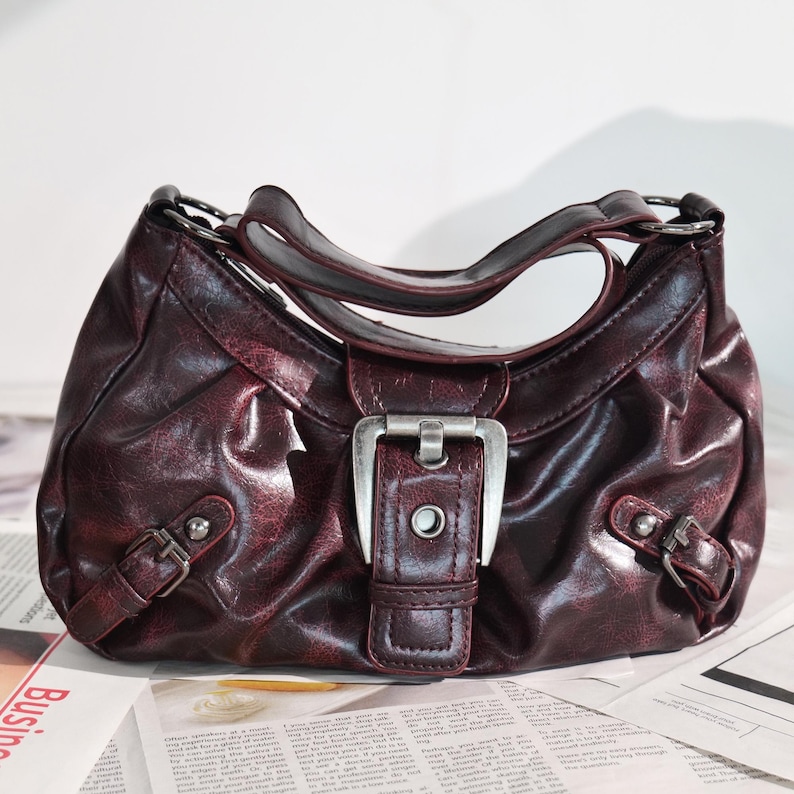 May include: A burgundy handbag with a silver buckle and accents. The bag has a curved handle and two smaller buckles on the sides. The bag is sitting on a newspaper.