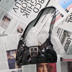 May include: A dark brown shoulder bag with a shiny finish, featuring a silver buckle and strap details. The bag is set against a backdrop of newspaper clippings, creating a vintage aesthetic.