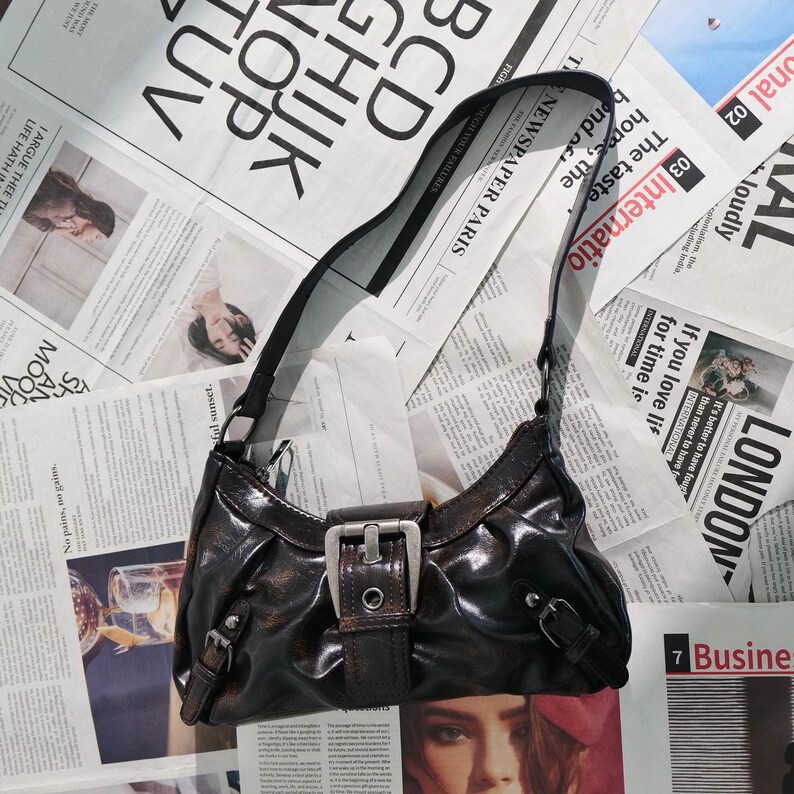 May include: A dark brown shoulder bag with a shiny finish, featuring a silver buckle and strap details. The bag is set against a backdrop of newspaper clippings, creating a vintage aesthetic.