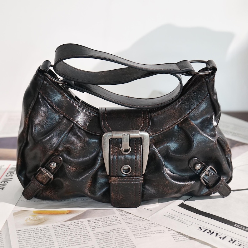 May include: A dark brown handbag with a curved shape and a single handle. The bag features a large silver buckle on the front and smaller buckles on the sides. The bag is made of a textured material and sits on a newspaper.