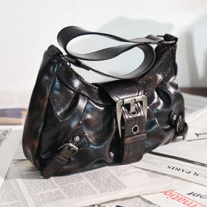 May include: A dark brown shoulder bag with a distressed finish. The bag features a curved handle, silver-colored buckles, and a flap closure. The bag is sitting on top of a newspaper.