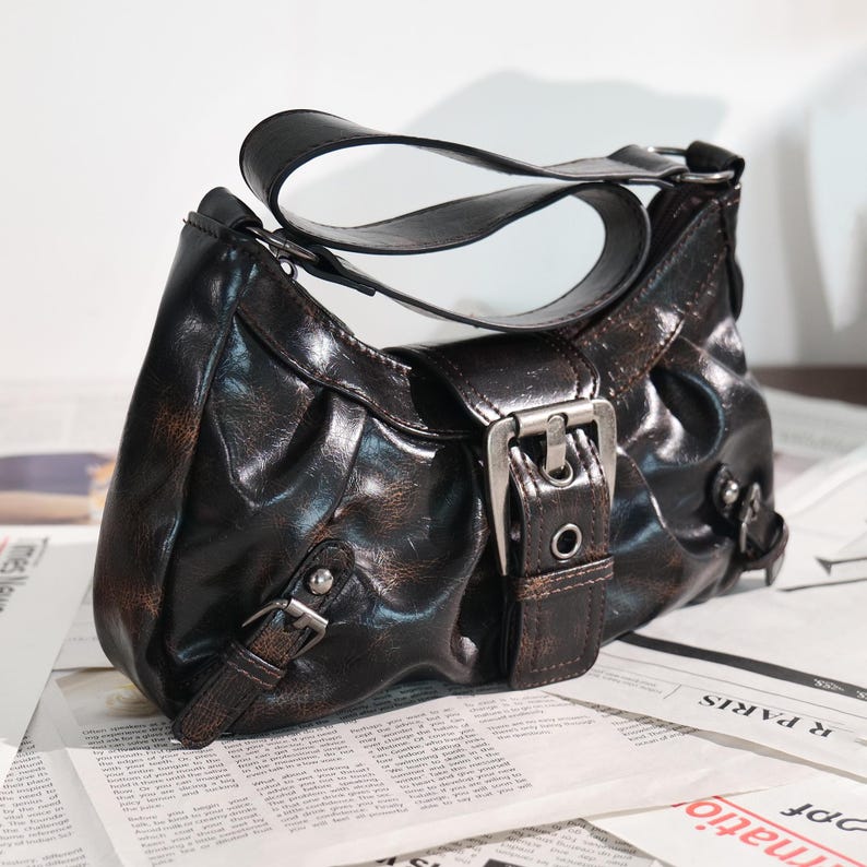 May include: A dark brown shoulder bag with a distressed finish. The bag features a curved handle, silver-colored buckles, and a flap closure. The bag is sitting on top of a newspaper.