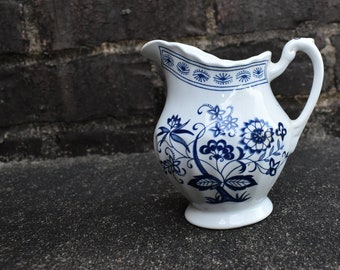 May include: White ceramic jug with a blue floral pattern. The jug features a curved handle and a scalloped rim with a blue border. The floral design includes stylized flowers and leaves. The jug is on a dark surface.