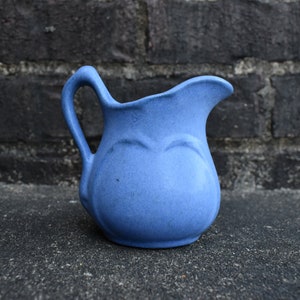 May include: A small, blue ceramic pitcher with a curved handle and a spout. The pitcher has a rounded body with a decorative embossed design. The background is a dark brick wall.