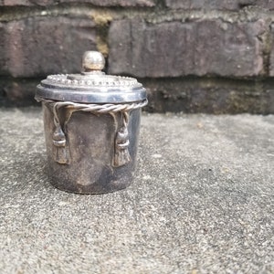 May include: A small, antique silver container with a lid. The container has a cylindrical shape with a decorative rope and tassel design. The lid is topped with a small, round knob. The container is placed on a concrete surface.