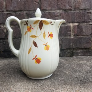 May include: Cream-colored ceramic pitcher with a lid and a curved handle. The pitcher is decorated with a floral pattern in shades of orange, yellow, and brown. The background is a brick wall.