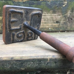 May include: A vintage, rectangular branding iron with the letters "EC" and the number "1981" embossed on its face. The iron is dark with a rusty appearance, and features a wooden handle.