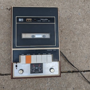 Könnte beinhalten: Vintage Realistic Stereo-Kassettendeck mit schwarzer und silberner Front. Das Deck hat eine eingelegte Kassette mit Tasten für Auswurf, Aufnahme, Wiedergabe, Stopp und Rücklauf. Das Gerät hat ein Netzkabel und liegt auf einer Betonoberfläche.