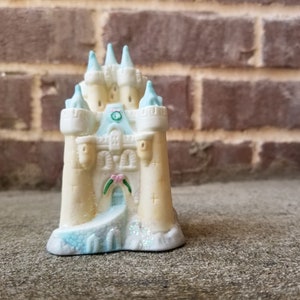 Puede incluir: Una pequeña figura decorativa de un castillo con cuerpo color crema y detalles en azul claro. El castillo presenta múltiples torres, puertas arqueadas y un pequeño puente. La base está decorada con purpurina. El fondo es una pared de ladrillo.