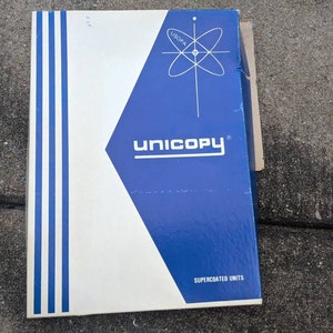 Może przedstawiać: Prostokątne pudełko z białym i niebieskim wzorem. Górna połowa jest niebieska z białym geometrycznym wzorem i napisem "UNICOPY" w kolorze białym. Dolna połowa jest biała z niebieskimi pionowymi paskami. Tekst "SUPERCOATED UNITS" znajduje się na dole.