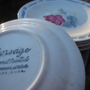 Puede incluir: Platos de cerámica blanca con diseño floral y detalles azules. La parte inferior de un plato dice "Corsage Hand Painted Dinnerware MADE IN U.S.A." Se ve una pila de platos, con enfoque en los detalles del patrón floral pintado a mano.