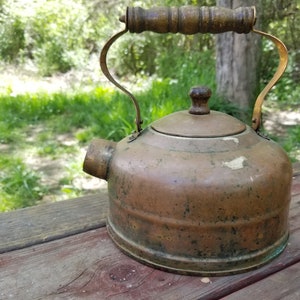 Könnte beinhalten: Vintage Kupferkessel mit Holzgriff und Messingakzenten. Der Kessel hat einen abgerundeten Körper, einen Ausguss und einen Deckel mit einem kleinen Holzknauf. Der Kessel steht auf einer Holzoberfläche mit verschwommenem grünem Hintergrund.