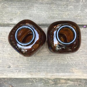 Puede incluir: Dos jarrones de cristal marrón con forma cuadrada y esquinas redondeadas. Cada jarrón tiene una abertura ancha con un borde blanco. Los jarrones están sobre una superficie de madera envejecida.