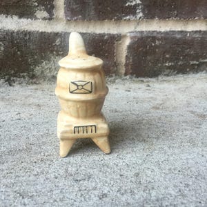 May include: A ceramic salt shaker in the shape of a small stove. The beige-colored shaker has a lid with a small handle and a black envelope design. It sits on a three-legged base with a black line design.
