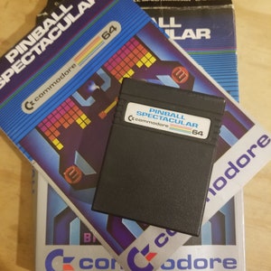 May include: A vintage Commodore 64 game cartridge and box set. The box features a blue and silver design with the title "Pinball Spectacular" and the Commodore logo. The cartridge is black with a label that reads "Pinball Spectacular 64".