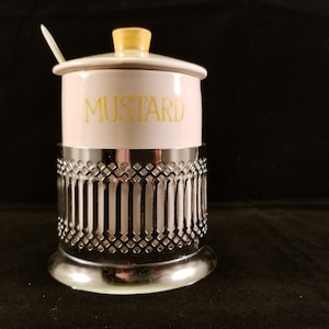 Puede incluir: Un tarro de mostaza vintage con cuerpo de cerámica blanca y tapa amarilla. La palabra "MUSTARD" está impresa en amarillo. Se encuentra en un soporte decorativo de metal plateado con una cuchara pequeña.