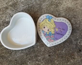 Puede incluir: Una caja de baratijas blanca en forma de corazón con tapa. La tapa presenta una niña de dibujos animados con cabello rubio, un lazo azul y el texto "You Are My Happiness". La caja está abierta, revelando su interior vacío.