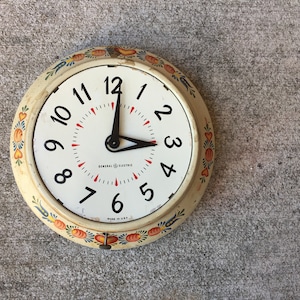 May include: Vintage General Electric wall clock with a cream-colored face and black numerals. The clock has a decorative floral border with red, orange, and blue accents. The hands point to 3:00. The text "MADE IN USA" is visible.