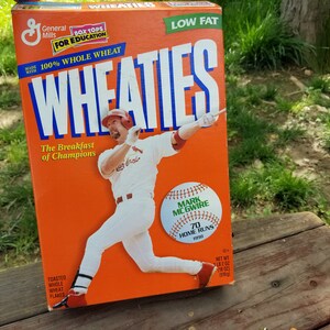 Könnte beinhalten: Eine orangefarbene Wheaties-Müsli-Schachtel mit einem Baseballspieler in weißer Uniform mit roten Akzenten. Die Schachtel enthält den Text "WHEATIES" in großen weißen Buchstaben und "The Breakfast of Champions."