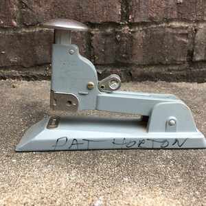 May include: A vintage gray stapler with a silver top and metal components. The name "PAT HORTON" is handwritten in black marker on the base. The stapler is set against a brick wall and concrete surface.