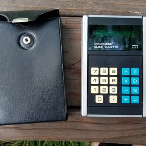 May include: A vintage Unisonic 888 Slide Rulette calculator with a black and silver design, featuring a numerical keypad and function buttons. It is accompanied by a black leatherette case with a snap closure. The calculator measures approximately 15 cm in length.