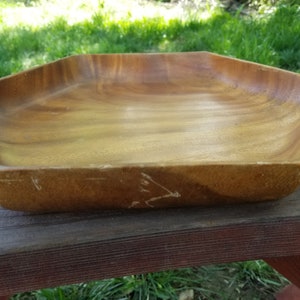 Puede incluir: Un cuenco de madera para servir con una forma geométrica única. El cuenco es de madera marrón claro y tiene una superficie lisa y pulida. Está sobre una mesa de picnic de madera.