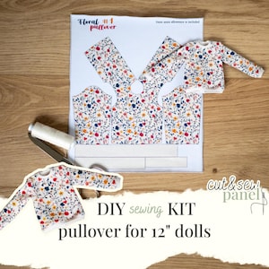 Könnte beinhalten: Ein DIY-Nähset für einen floralen Pullover für 30 cm Puppen. Das Set enthält Stoffstücke, eine Garnrolle und eine Schere. Der Stoff hat einen weißen Hintergrund mit einem bunten Blumenmuster. Der Text "Floral #1 pullover" ist sichtbar.