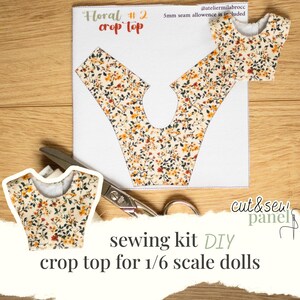 Puede incluir: Un kit de costura DIY para un top corto para muñecas a escala 1/6. La tela floral presenta un fondo crema con pequeños estampados florales naranjas, rojos y verdes. El kit incluye el patrón, una pieza recortada y un top corto terminado. También se ven unas tijeras.