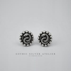 Op de afbeelding: Een paar ronde zilveren oorknopjes met een ontwerp van een opgerolde slang in het midden, omringd door een rand van kleine zilveren bolletjes. De tekst GOTHIC SILVER ATELIER 925 STERLING SILVER staat onder de oorbellen op een lichtgrijze achtergrond.