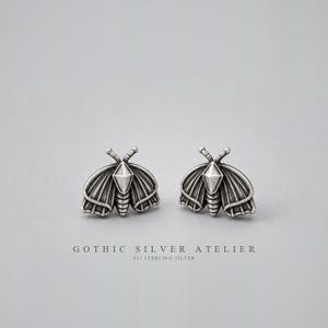 May include: A pair of sterling silver stud earrings shaped like moths with detailed wing patterns and a central diamond-shaped accent. The text GOTHIC SILVER ATELIER 925 STERLING SILVER is printed below the earrings on a plain background.