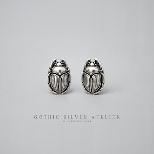 May include: A pair of silver stud earrings shaped like scarab beetles with detailed ridged wings and a polished finish. The text GOTHIC SILVER ATELIER 925 STERLING SILVER is printed below the earrings on a plain light gray background.