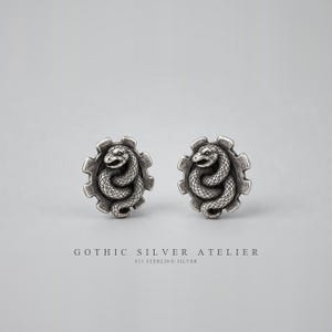 May include: A pair of sterling silver stud earrings featuring a coiled snake design centered within a gear-shaped frame. The metal has an oxidized finish to highlight the detailed scales of the serpent and the mechanical texture of the gear.