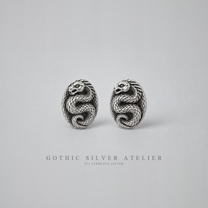 May include: A pair of oval-shaped sterling silver stud earrings featuring a coiled snake design with detailed scales and a defined head. Below the earrings, the text reads GOTHIC SILVER ATELIER 925 STERLING SILVER in a clean, sans-serif font.