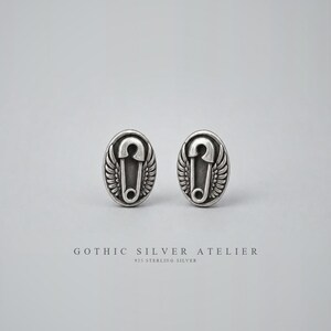 Op de afbeelding: Een paar ovale zilveren oorbellen met een centraal veiligheidsspeldmotief geflankeerd door gedetailleerde vleugels. De tekst GOTHIC SILVER ATELIER 925 STERLING SILVER staat onder de oorbellen. Het metaal heeft een donkere, geoxideerde afwerking.