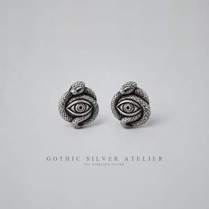 Puede incluir: Un par de pendientes de plata con una serpiente detallada enroscada alrededor de un diseño de ojo central. El metal tiene un acabado oxidado para resaltar las escamas y los detalles del iris. El texto GOTHIC SILVER ATELIER 925 STERLING SILVER aparece debajo.