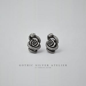 Könnte beinhalten: Ein Paar silberne Ohrstecker mit einer detaillierten Rose, die von einer gewundenen Schlange umschlungen wird. Das Metall hat ein oxidiertes Finish, um die texturierten Schuppen der Schlange und die Blütenblätter der Rose hervorzuheben. Der Text lautet GOTHIC SILVER ATELIER 925 STERLING SILVER.
