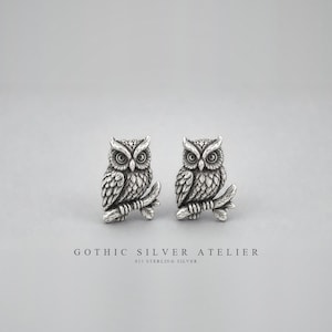 May include: A pair of sterling silver stud earrings shaped like owls perched on branches. The metal has an oxidised finish to highlight the detailed feather textures and large eyes. The text GOTHIC SILVER ATELIER 925 STERLING SILVER is below.