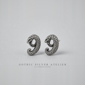 Könnte beinhalten: Ein Paar silberne Ohrstecker in Form von gewundenen Schlangen mit detaillierter Schuppenstruktur und oxidierten Akzenten. Der Text GOTHIC SILVER ATELIER 925 STERLING SILVER ist unter den Ohrringen auf einem hellgrauen Hintergrund gedruckt.