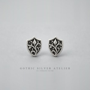 Op de afbeelding: Een paar zilveren oorknopjes in de vorm van een schild, voorzien van een reliëfpatroon van ranken en bladeren met geoxideerde details. De tekst GOTHIC SILVER ATELIER 925 STERLING SILVER staat onder de oorbellen op de ondergrond.