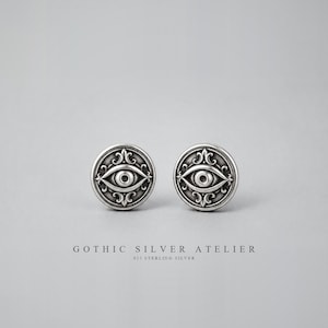 May include: A pair of round silver stud earrings featuring an embossed eye design surrounded by intricate scrollwork patterns. The text GOTHIC SILVER ATELIER 925 STERLING SILVER is printed below the earrings on the plain light grey background.