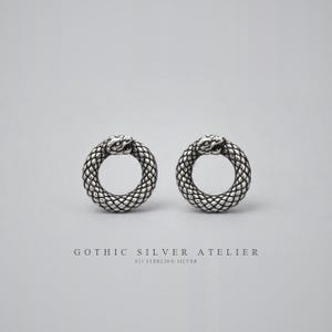 Può includere: Un paio di orecchini a lobo circolari in argento sterling, ognuno raffigurante un serpente che morde la propria coda in un design ouroboros con squame testurizzate. Il testo GOTHIC SILVER ATELIER 925 STERLING SILVER appare qui sotto.