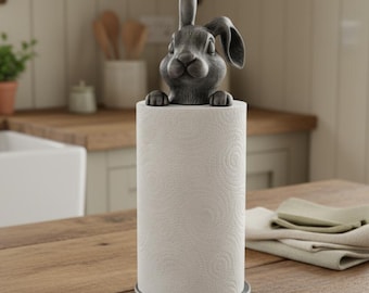 May include: A gray bunny-shaped paper towel holder. The bunny is perched atop a roll of white paper towels, with its ears and head visible. The holder has a circular base and is set on a wooden surface. Kitchen decor.