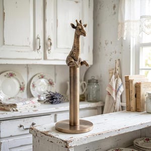 May include: A bronze-toned metal paper towel holder featuring a sculpted giraffe figure at the top. The stand consists of a circular base, a vertical cylindrical rod, and a detailed giraffe head and front legs resting on the top of the rod.