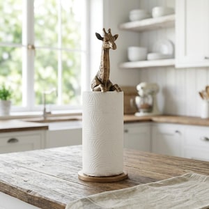 May include: A paper towel holder featuring a bronze-toned giraffe figurine topper. The holder consists of a circular base and a vertical rod, with a roll of white paper towels positioned around the rod. The giraffe is depicted in a seated position.