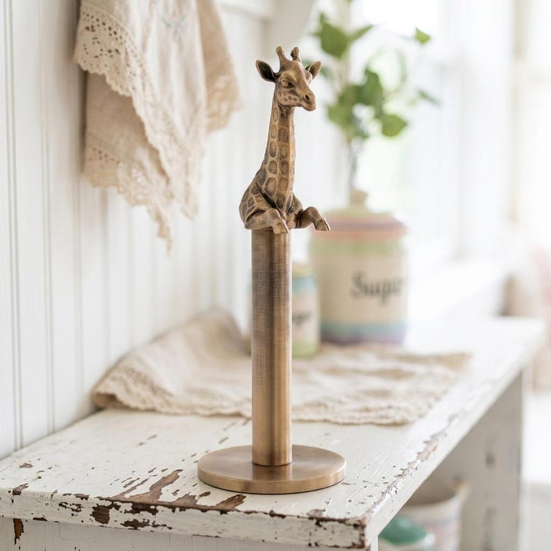 May include: A vertical paper towel holder made of brushed bronze-toned metal. The base is a flat circular disc supporting a central cylindrical rod. At the top of the rod sits a detailed, three-dimensional figurine of a giraffe head and torso.