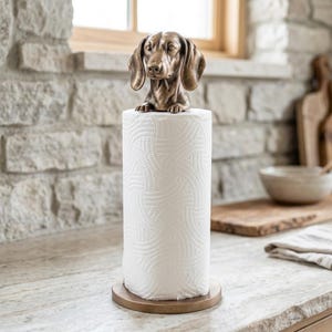 May include: A bronze-toned metal paper towel holder featuring a sculpted dachshund dog head and front paws at the top. The holder has a circular base and holds a standard roll of white paper towels with a textured, embossed pattern on the surface.