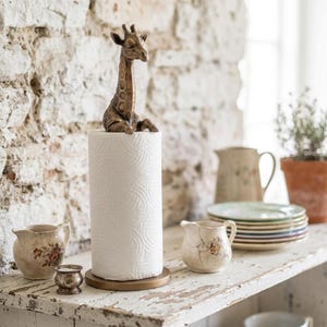May include: A paper towel holder featuring a bronze-toned giraffe figurine perched atop a cylindrical roll of white paper towels. The holder has a circular base and a vertical rod that supports the paper towel roll, with the giraffe head at the top.