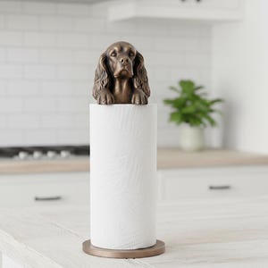 May include: A bronze-colored dog-shaped paper towel holder. The dog figure is perched atop a roll of white paper towels. The holder has a round base and is designed for kitchen use.